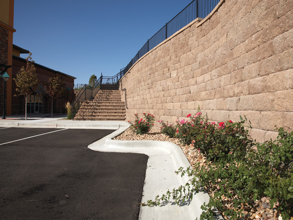 Parking lot with curved Diamond Pro stone cut concrete block retaining wall and  stone staircase, topped with a black metal fence 