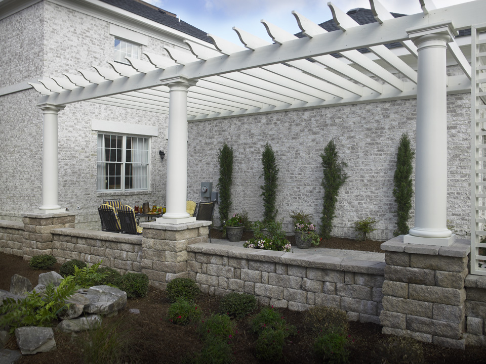 Grey brick building flanked by a stone paver walkway with Highland Stone concrete block walls and a white wooden pergola