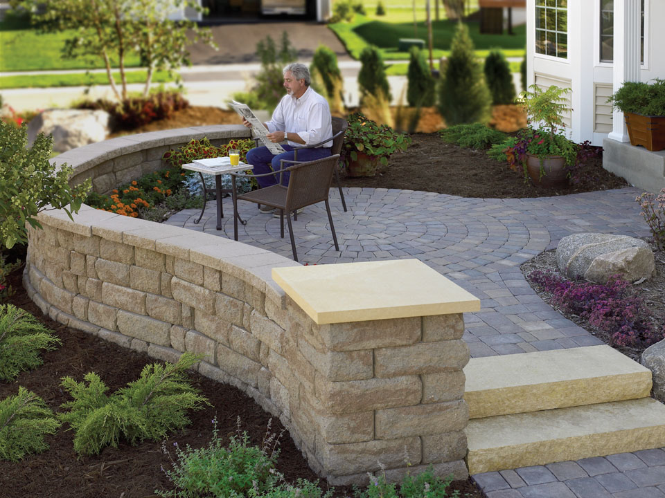 Man reading a newspaper on a stone paver patio with a curved Highland Stone concrete block wall and concrete step units