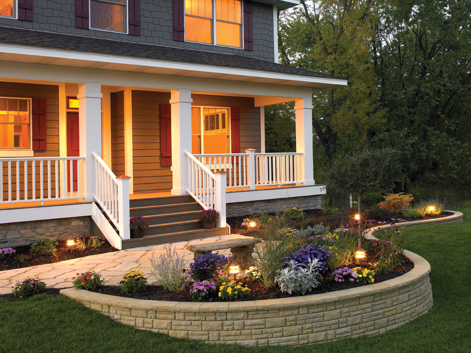 Front porch of a gray house with a paving stone walkway and curved raised Natural Impressions Ashlarstone concrete block retaining wall planter. 